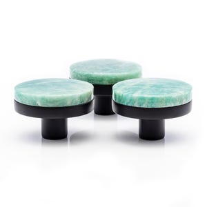May include: Three cabinet knobs with light green, circular tops and black bases. The knobs have a marbled effect, with variations in the green colour. The bases are cylindrical and have a matte finish. The knobs are set against a white background.