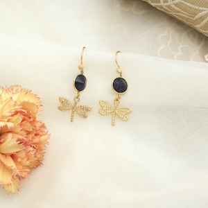 Może przedstawiać: Para złotych kolczyków z motywem ważki. Każdy kolczyk ma ciemnoniebieski, owalny kamień zawieszony na złotym haczyku. Poniżej kamienia wisi szczegółowy urok ważki, tworząc delikatny i inspirowany naturą design.
