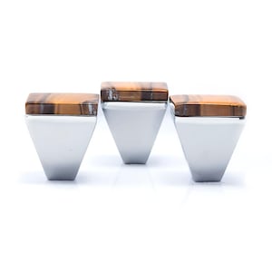 May include: Three silver rings with a geometric design. Each ring features a square-shaped, tiger's eye stone set atop a silver band. The rings are displayed against a white background, highlighting their polished appearance.