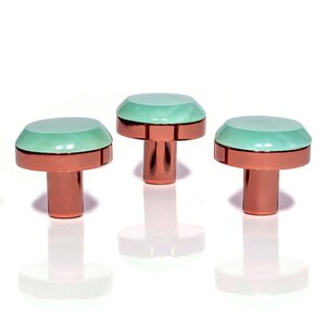 Op de afbeelding: Drie decoratieve kastknoppen met een koperkleurige basis en een mintgroene, facetgeslepen bovenkant. De knoppen zijn in een rij gerangschikt tegen een witte achtergrond, met hun reflecties eronder zichtbaar. De knoppen hebben een modern, geometrisch ontwerp.