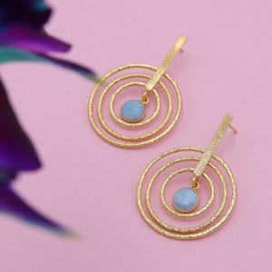 Może przedstawiać: Para złotych kolczyków z teksturowanym wykończeniem. Każdy kolczyk ma trzy koncentryczne okręgi i prostokątny pasek. Jasnoniebieski kamień szlachetny wisi ze środka okręgów. Kolczyki są na różowym tle.