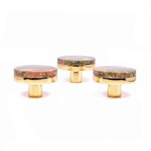 Könnte beinhalten: Drei dekorative Schrankknöpfe. Jeder Knopf hat eine runde, gesprenkelte Oberseite in Rot-, Grün- und Brauntönen, die auf einer goldfarbenen Basis angebracht ist. Die Knöpfe werden vor einem weißen Hintergrund präsentiert.