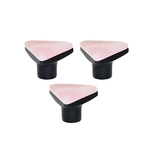 Pode incluir: Três puxadores decorativos para armários. Cada puxador apresenta um topo triangular rosa marmoreado com uma borda preta, colocado sobre uma base cilíndrica preta. Estes puxadores são projetados para móveis e decoração de casa.