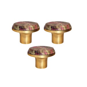 May include: Three cabinet knobs with a decorative design. Each knob has a round top made of multi-coloured stone with pink, brown, and gold tones, set on a gold-coloured metal base. Ideal for furniture and home decor.