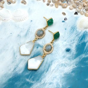 May include: Gold-coloured earrings featuring a green square stud, a grey gemstone in a round setting, and a white geometric pendant. The earrings are presented on a blue and white marbled surface, with seashells and small stones in the background.
