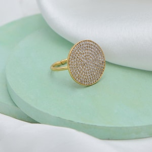 May include: A gold-toned ring with a large, circular face covered in small, sparkling stones. The ring is set against a light green surface with a white fabric in the background. The ring's band is thin and simple.