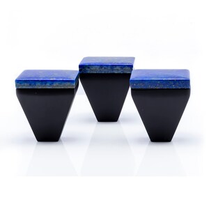 Puede incluir: Tres cajas geométricas negras con tapas cuadradas moteadas azules. Las cajas tienen forma triangular, con la base más ancha que la parte superior. Las tapas azules tienen un aspecto moteado, que contrasta con los cuerpos negros sólidos. Las cajas están dispuestas sobre una superficie blanca.