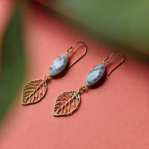 May include: A pair of gold-coloured drop earrings. Each earring has a light blue and white oval stone and a gold-coloured leaf charm. The earrings are set against a pink and green background.