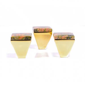 Op de afbeelding: Drie decoratieve dozen met een geometrisch ontwerp. Elke doos heeft een goudkleurige, trapeziumvormige basis en een vierkante bovenkant van een gespikkelde, roodgroene steen. De dozen zijn op een witte achtergrond geplaatst.