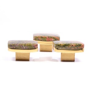 Könnte beinhalten: Drei quadratische Schrankknöpfe mit einer Oberseite aus Naturstein in Grün- und Rottönen. Die Knöpfe haben eine goldfarbene Basis und eine quadratische Form. Diese dekorativen Knöpfe sind ideal, um Möbeln einen Hauch von natürlicher Eleganz zu verleihen.