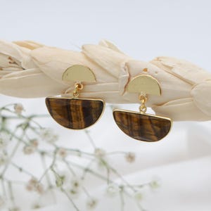 May include: A pair of gold-toned earrings with a half-moon design. Each earring features a gold half-circle at the top, connected to a half-moon shaped stone with brown and gold tiger-eye patterns. The earrings are displayed on a white background.