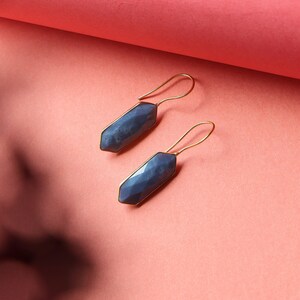 Może przedstawiać: Para kolczyków z niebieskim kamieniem szlachetnym ze złotymi haczykami. Kolczyki mają geometryczny, fasetowany design i są umieszczone na różowym tle. Modny dodatek.