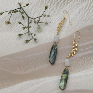 Pode incluir: Um par de brincos dourados com um design de folha. Cada brinco apresenta uma pedra azul-esverdeada iridescente em forma de gota, uma pequena pedra branca e um gancho dourado. Os brincos são exibidos em uma superfície texturizada esbranquiçada.