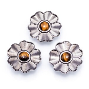 May include: Three silver-coloured flower-shaped buttons with a central, round, tiger's eye stone. The petals have a textured, ridged design. The buttons are arranged on a white background.