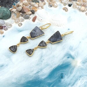 May include: Gold-coloured earrings featuring three grey, geometric stones. The earrings have a hook fastening and are set against a blue and white background with seashells and pebbles.