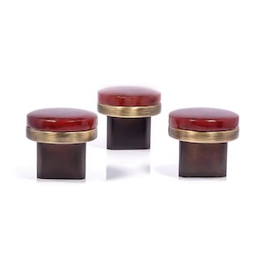 May include: Three cabinet knobs with a dark red, glossy top and a brass-coloured band. The base is a dark brown, rectangular shape. The knobs are arranged in a row against a white background.