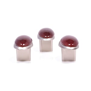 Może przedstawiać: Trzy gałki do szafek z prostokątną podstawą i zaokrąglonym, czerwonobrązowym kamiennym blatem. Kamień osadzony jest w srebrnym metalowym pierścieniu. Gałki są prezentowane na białym tle.