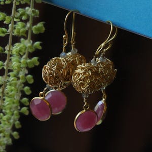 May include: Gold-coloured wire-wrapped earrings with a woven ball design, featuring pink gemstone drops. The earrings have a delicate, handcrafted appearance, with intricate wirework and the vibrant colour of the gemstones.
