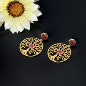 May include: Gold-coloured earrings with a tree of life design. Each earring has a circular, openwork pendant with a red gemstone centre and a smaller red gemstone at the top. A white and yellow flower is in the background.