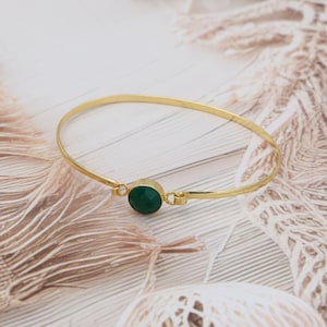 May include: A gold-coloured bangle bracelet featuring a round, faceted, dark green gemstone. The bracelet has a simple, elegant design, with the gemstone set in a gold bezel.
