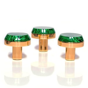 May include: Three decorative bottle stoppers. Each stopper has a gold-coloured base and a top with a green, marbled design. The stoppers are arranged in a row against a white background, with their reflections visible below.