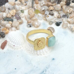 Puede incluir: Un anillo dorado con una piedra preciosa cuadrada de color azul claro y un engaste cuadrado decorativo. El anillo está sobre una superficie blanca con pequeños guijarros y conchas marinas.