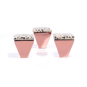 May include: Three rose gold cabinet knobs with square speckled stone tops. The knobs have a geometric, trapezoid shape. The speckled stone has a mix of black, grey, and white colours. The knobs are set against a white background.