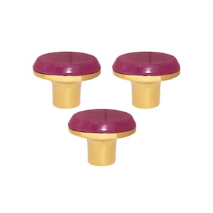 Può includere: Tre pomelli decorativi per mobili. Ogni pomello presenta una parte superiore rotonda color magenta scuro e una base dorata. I pomelli sono disposti a triangolo. Ogni pomello ha un diametro di circa 3 cm.