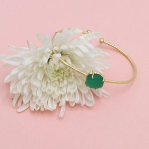 Pode incluir: Uma pulseira de punho dourada com uma vibrante pedra preciosa verde. A pulseira é aberta, com um design liso e curvo e uma única pedra verde de formato irregular. A pulseira está sobre uma flor branca e um fundo rosa suave.