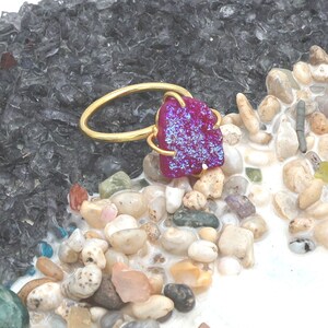 May include: A gold-colored ring featuring a vibrant, textured, purple gemstone. The gemstone has a sparkling, crystalline appearance. The ring is set against a backdrop of small, smooth stones and a dark, textured surface.