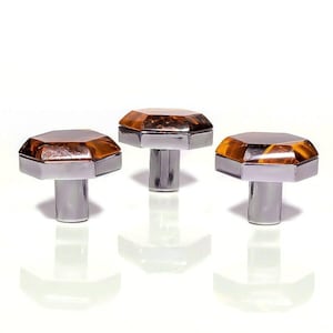 May include: Three cabinet knobs with an amber-coloured, faceted top and a silver-coloured base. The knobs have a geometric design and are arranged in a row against a white background. Suitable for furniture upgrades.