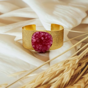 May include: A gold cuff bracelet with a textured finish, featuring a large, oval-shaped, fuchsia crystal. The bracelet is set against a soft, off-white background with dried wheat stalks.