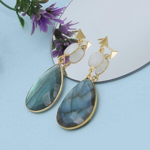 Pode incluir: Brincos dourados com pedras de labradorita em forma de gota, exibindo iridescência azul-esverdeada. O design incorpora pedras ovais brancas e pequenos triângulos dourados. Apresentados em uma superfície reflexiva.