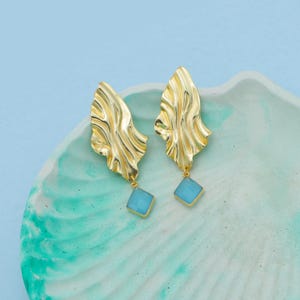 May include: A pair of gold-colored earrings with a textured, leaf-like design. Each earring features a light blue, square-shaped gemstone pendant. The earrings are displayed on a light blue and green surface against a light blue background.