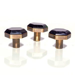 Könnte beinhalten: Drei dekorative Schrankknöpfe. Jeder Knopf hat eine facettierte, dunkelblaue Oberseite mit Goldflocken, ein goldfarbenes Band und einen zylindrischen Messingstiel. Die Knöpfe sind auf einer weißen Oberfläche angeordnet.