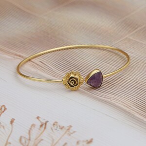 Op de afbeelding: Goudkleurige armband met een bloemmotief en een driehoekige paarse edelsteen. De armband heeft een delicaat, open ontwerp. De edelsteen is gezet in een gouden bezel. Een stijlvol sieraad voor dagelijks gebruik.