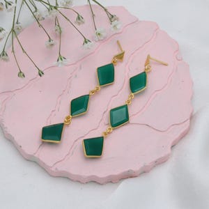 Könnte beinhalten: Ein Paar goldfarbene Ohrhänger mit smaragdgrünen, rautenförmigen Edelsteinen. Die Ohrringe werden auf einer rosa Oberfläche mit weißen Blumen im Hintergrund präsentiert. Die Edelsteine sind durch kleine goldene Glieder verbunden.