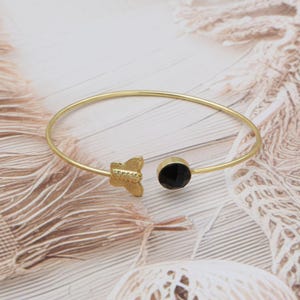 May include: A gold-coloured bangle bracelet featuring a butterfly motif and a black gemstone. The butterfly is on one end, and the gemstone is on the other. The bracelet is on a light-coloured surface.