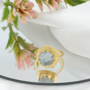 Può includere: Un anello color oro con una pietra preziosa azzurra. L'anello ha un'incastonatura circolare strutturata e una fascia semplice. L'anello è esposto su una superficie riflettente, con fiori rosa sullo sfondo. Design elegante.