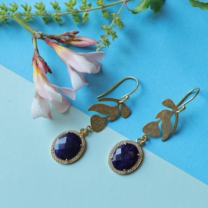 May include: A pair of gold-colored earrings with a leaf design and a dark blue, oval-shaped stone. The stone is surrounded by small, clear crystals. The earrings are displayed on a light blue and blue background with pink flowers.