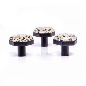 May include: Three cabinet knobs with a black and white speckled pattern on a black base. The knobs have an octagonal shape and a cylindrical stem. The knobs are set against a white background. The knobs are suitable for cabinets.
