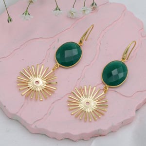 Peut inclure: Paire de boucles d'oreilles dorées avec une pierre verte ovale et un motif de soleil. Les boucles d'oreilles sont présentées sur une surface rose avec des fleurs blanches en arrière-plan. Fermeture à crochet.