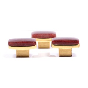 Peut inclure: Trois boutons de meuble avec des dessus rectangulaires brun-rouge et des bases dorées. Les boutons sont disposés en rangée, chacun avec une finition polie. Le matériel est conçu pour les meubles et la décoration intérieure.
