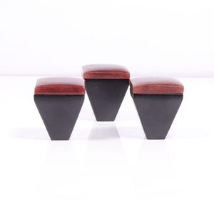 May include: Three contemporary stools. Each stool features a dark grey, trapezoid-shaped base and a reddish-brown, rectangular seat. The stools are arranged in a row against a white backdrop.