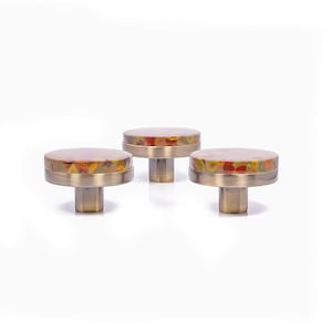Op de afbeelding: Drie decoratieve kastknoppen met een messing afwerking en kleurrijke, gespikkelde bovenkanten. De knoppen hebben een ronde vorm en een korte, cilindrische steel. De bovenkanten hebben een mix van rode, gele en groene tinten.