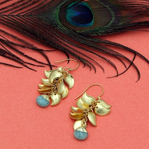 Peut inclure: Boucles d'oreilles pendantes dorées avec un motif de feuilles et une pierre précieuse bleu clair en bas. Les boucles d'oreilles sont sur un fond rouge avec une plume de paon en arrière-plan. Fermeture à crochet.