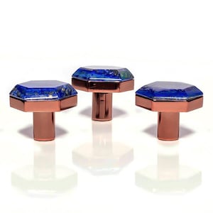 Op de afbeelding: Drie decoratieve kastknoppen met een geometrisch ontwerp. Elke knop heeft een gefacetteerde, diepblauwe edelsteen top met koperkleurige metalen accenten en een cilindrische basis. De knoppen zijn gerangschikt op een wit oppervlak.