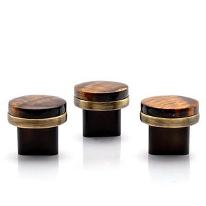 May include: Three bottle stoppers with a dark brown, rectangular base and a circular, wood-grain top. A brass-coloured band encircles the base of the wooden top. The design is elegant and would suit a variety of bottles.