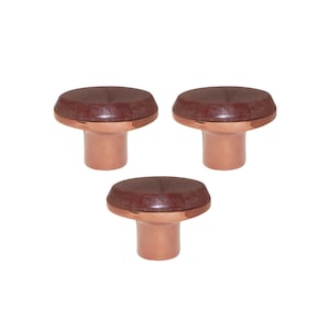 Pode incluir: Três puxadores de armário cor de cobre com um topo circular castanho escuro. Os puxadores têm formato de cogumelo, com uma base cilíndrica curta e um topo mais largo e arredondado. Os puxadores estão espaçados uniformemente sobre um fundo branco.