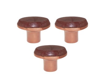 May include: Three copper-colored cabinet knobs with a dark brown, circular top. The knobs have a mushroom-like shape, with a short, cylindrical base and a wider, rounded top. The knobs are evenly spaced against a white background.